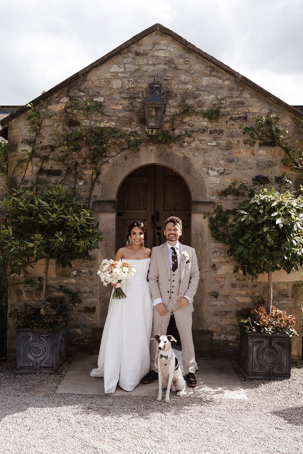 Couple and their white dog pose in front of a rustic stone chapel with an arched wooden door.