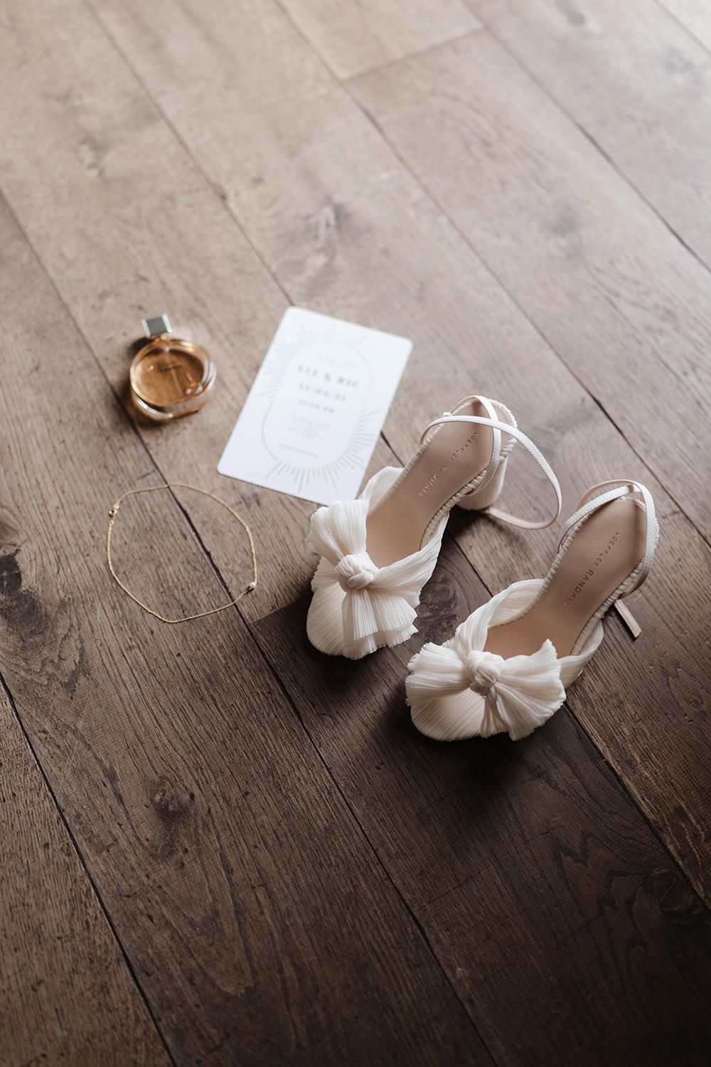 Elegant white bridal shoes with bow detail and wedding rings displayed on rustic wooden floor.