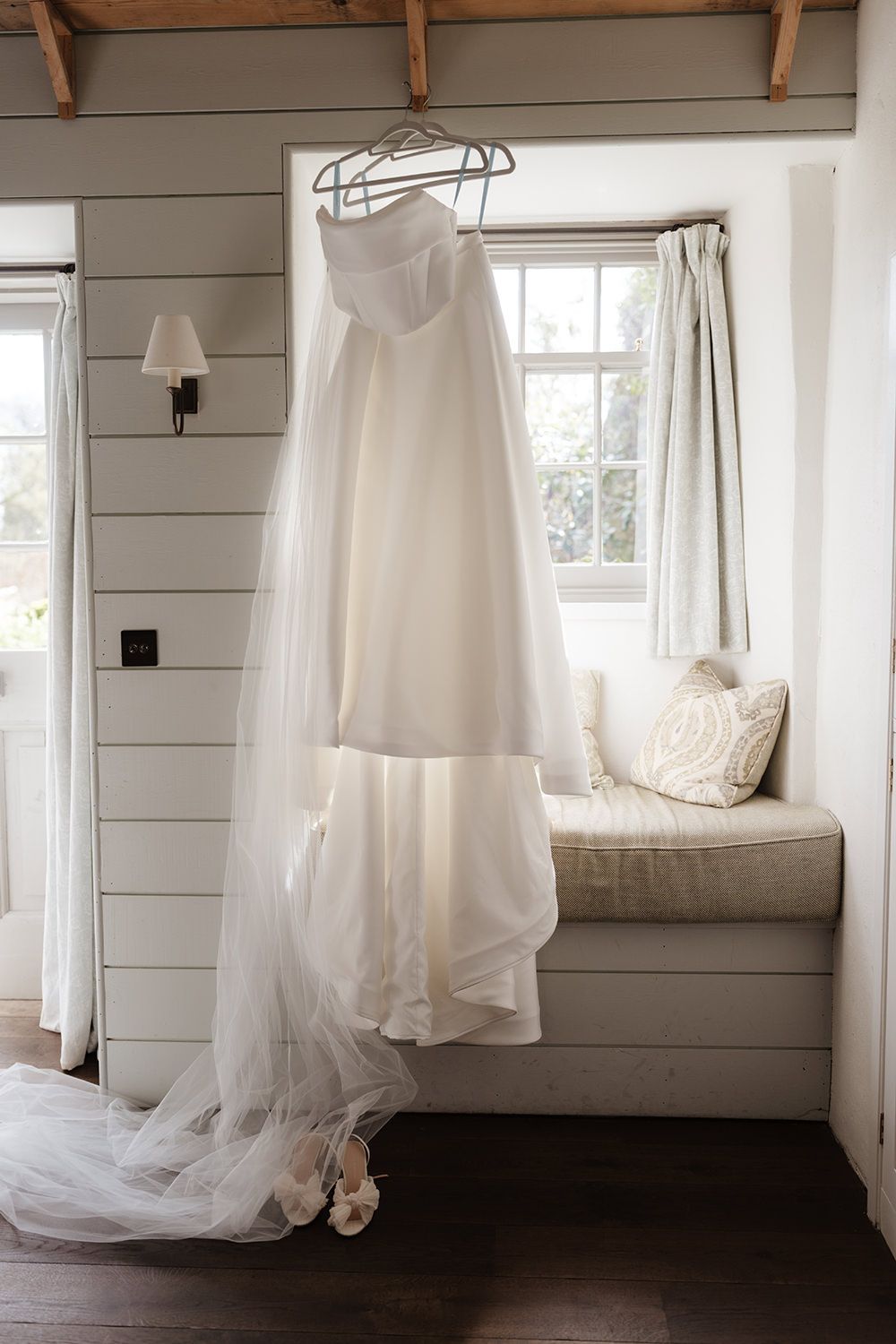 A white wedding dress hangs elegantly by a window nook with natural light streaming in, highlighting its flowing train and details.