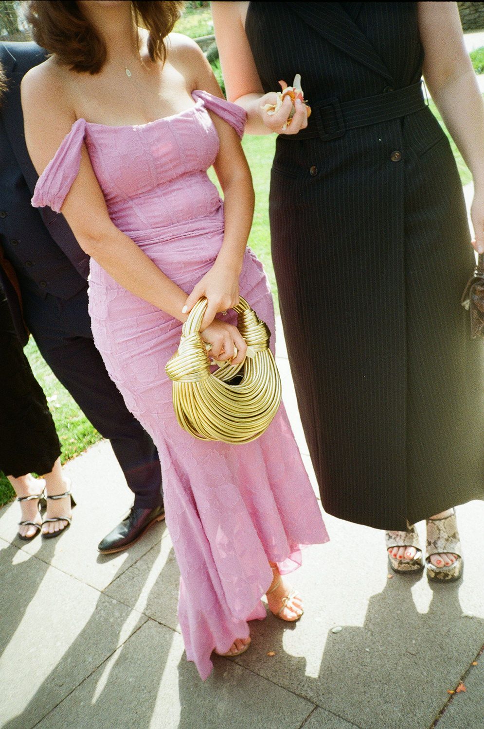 Woman in pink mermaid gown holds gold metallic handbag at formal outdoor event.