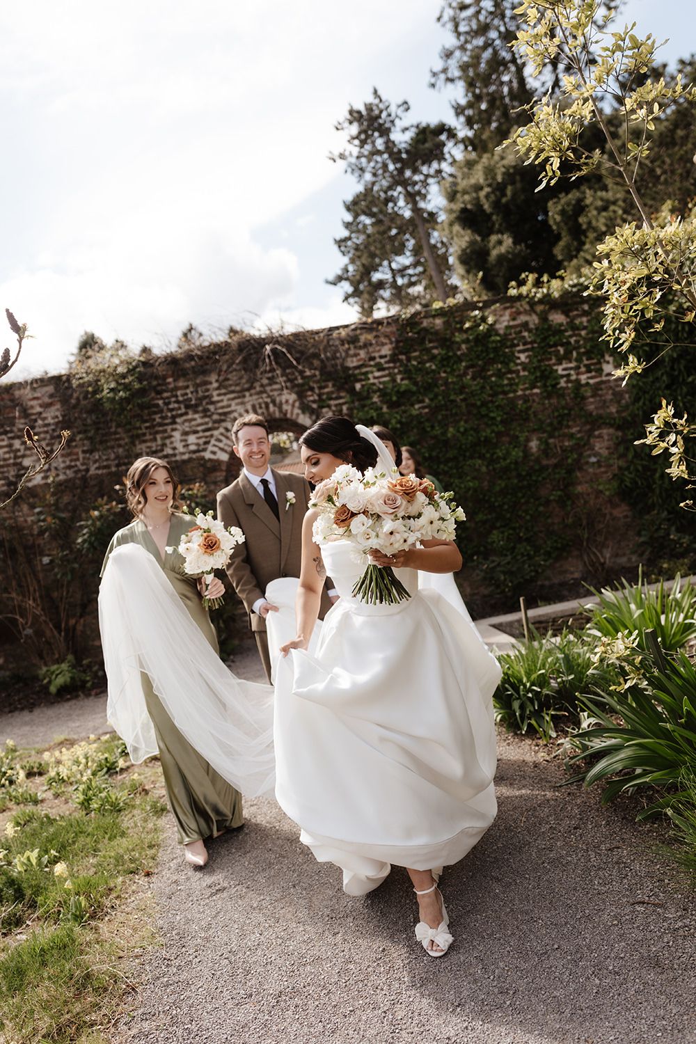 Wedding party walking along garden path by stone wall as bridesmaids help carry bride's flowing white gown train.