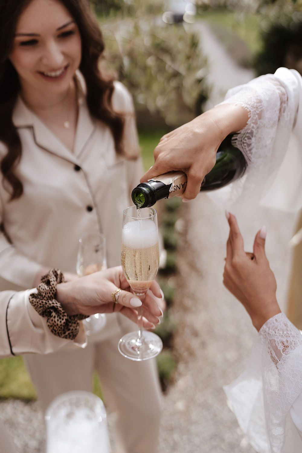 Hands pouring sparkling champagne into elegant fluted glasses during an outdoor celebration in lace sleeves and silk.