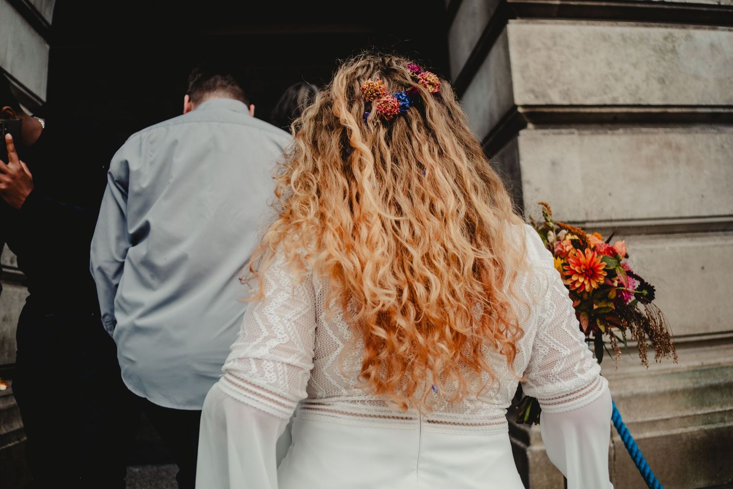 Bride with long curly blonde hair in white dress standing behind someone wearing gray at a stone building entrance.