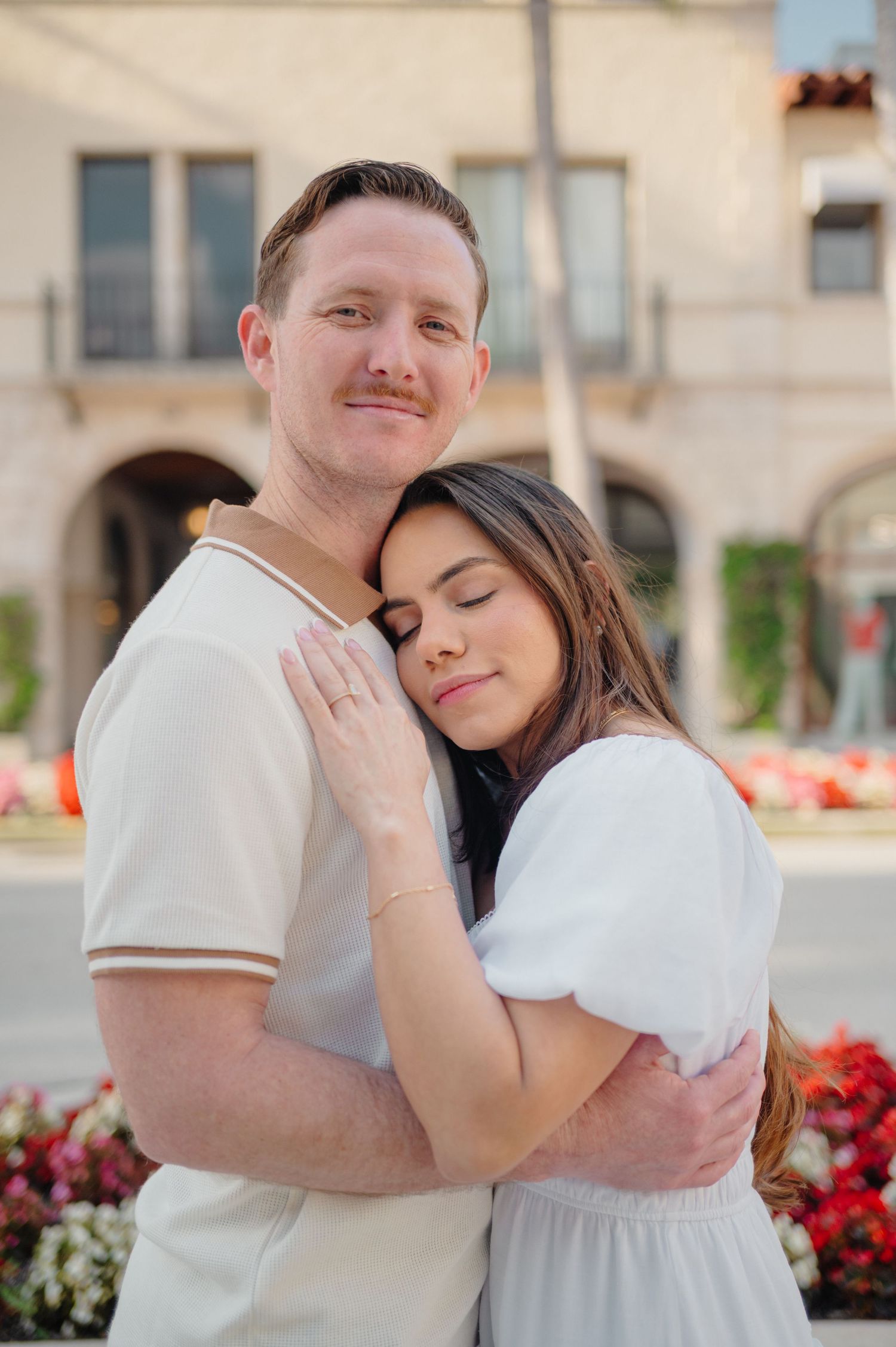 A romantic couple in white outfits embracing outdoors with red flowers and arched architecture in the background.