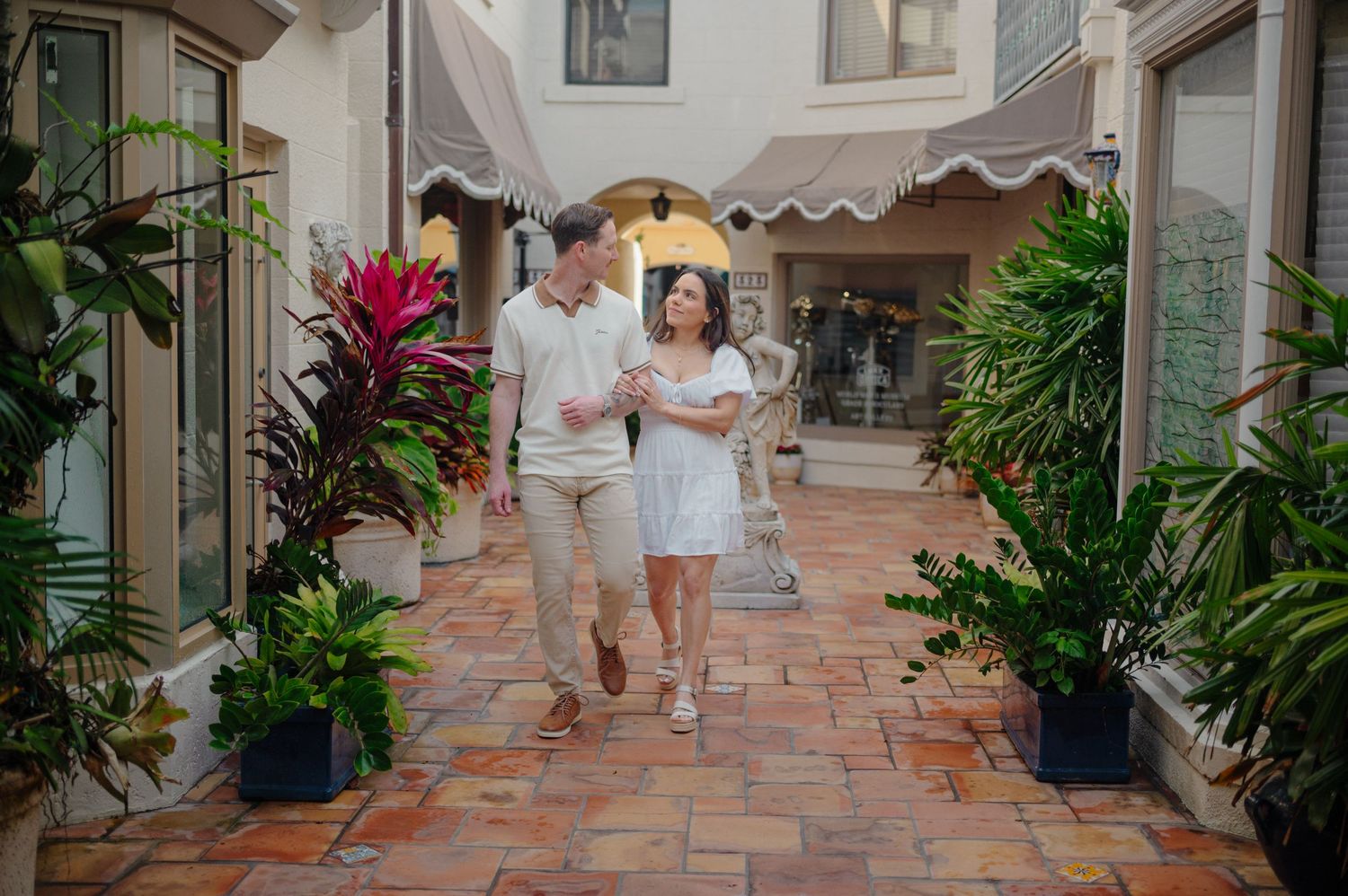 Two people walk down a scenic covered walkway lined with tropical plants and terracotta tiles.