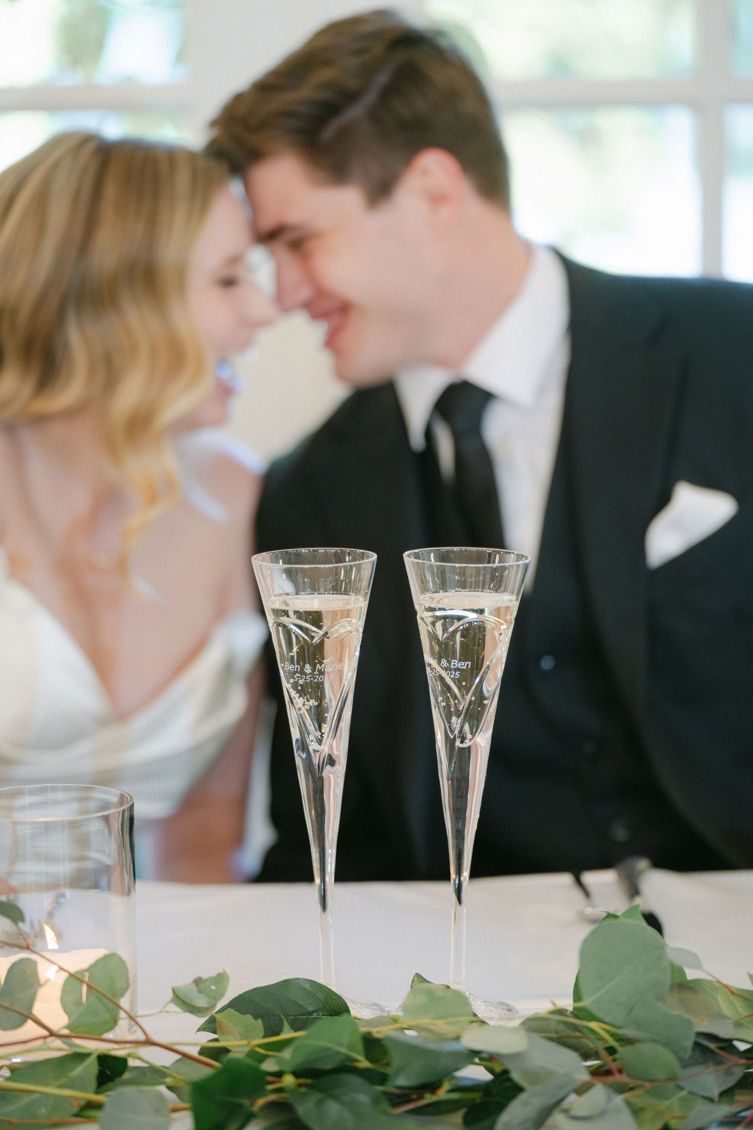 Two champagne flutes sit on a table decorated with eucalyptus leaves while a couple shares an intimate moment behind them.