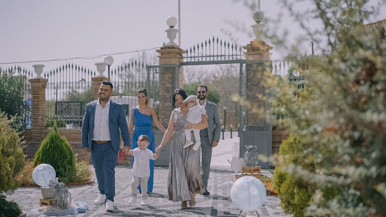 Family walking along decorated garden path with ornate gates and white balloons.