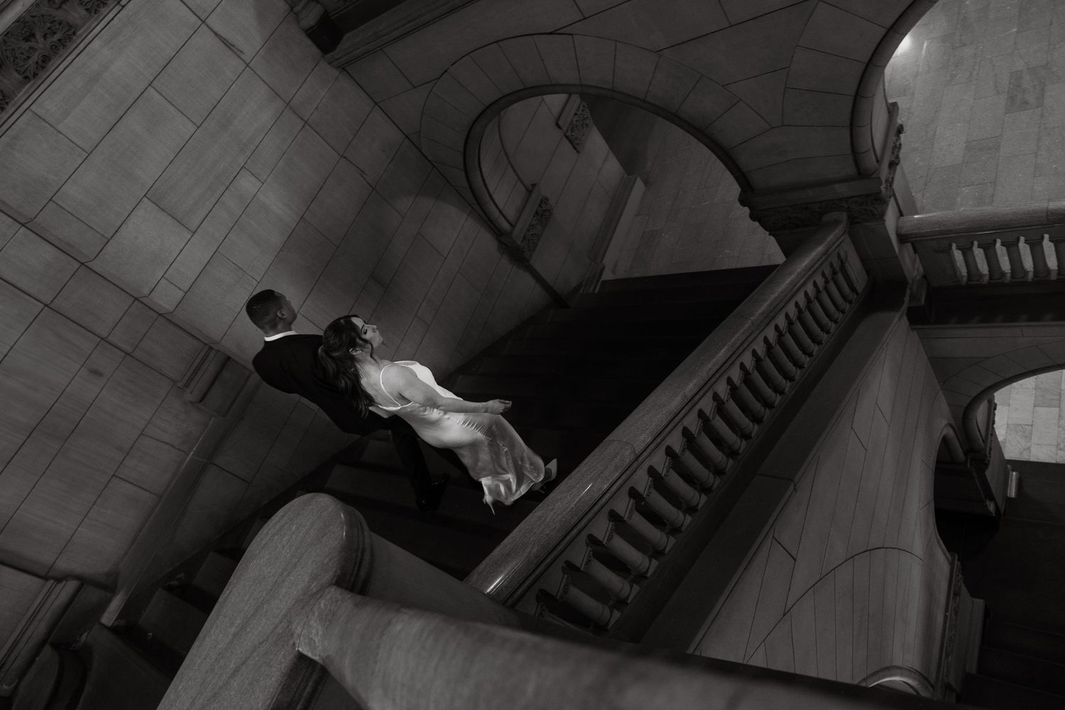 A person in formal attire stands at the top of an elegant marble staircase in a grand architectural setting.
