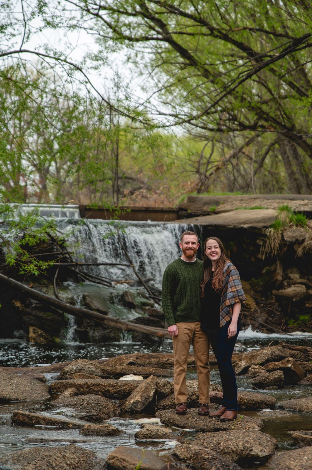 A couple stands on rocky terrain in front of a cascading waterfall surrounded by lush green trees.