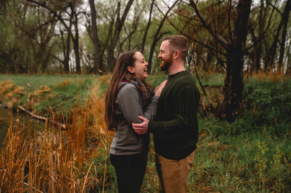 A couple shares an intimate moment while standing in tall autumn grass near a wooded area.