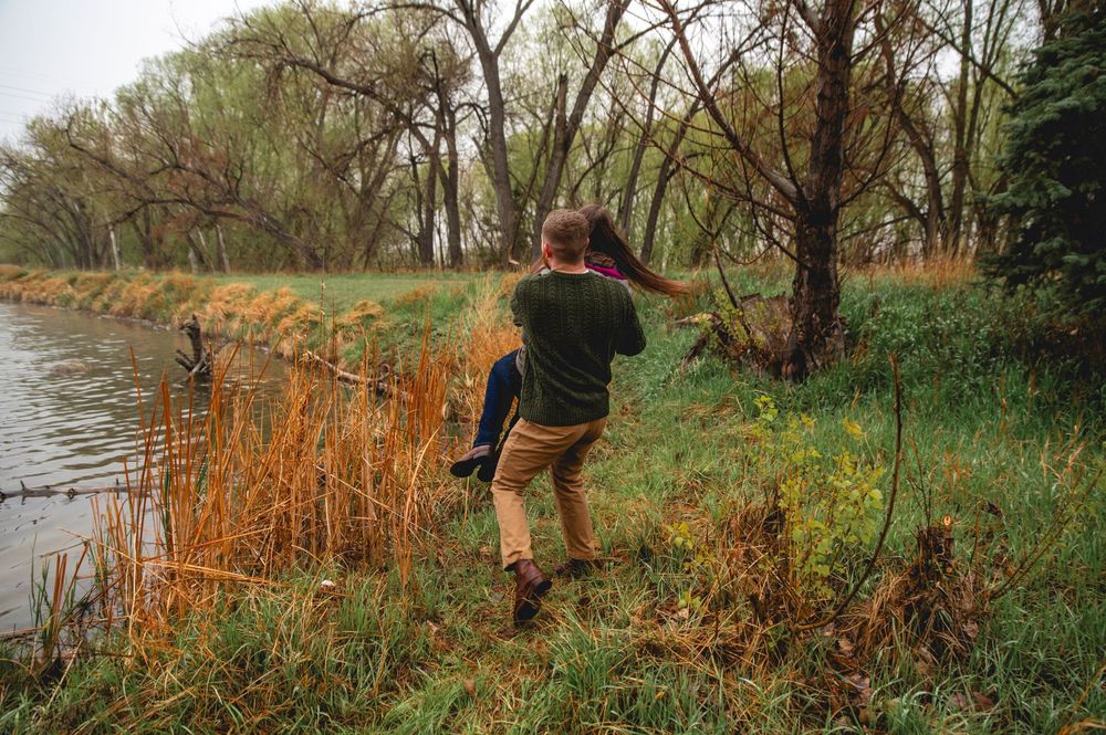 Someone takes a photo near a river bank with tall grass and autumn trees in the background.