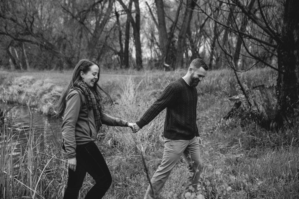 A couple holds hands while walking together through a wooded area in black and white photography.