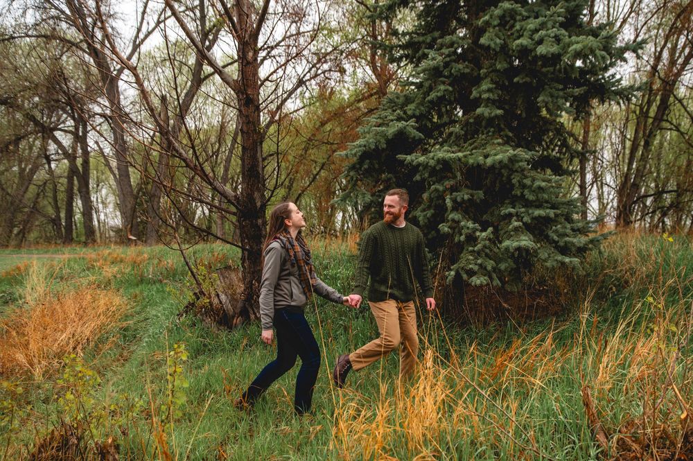 Two people walk through a scenic wooded area with tall grass and evergreen trees during autumn.
