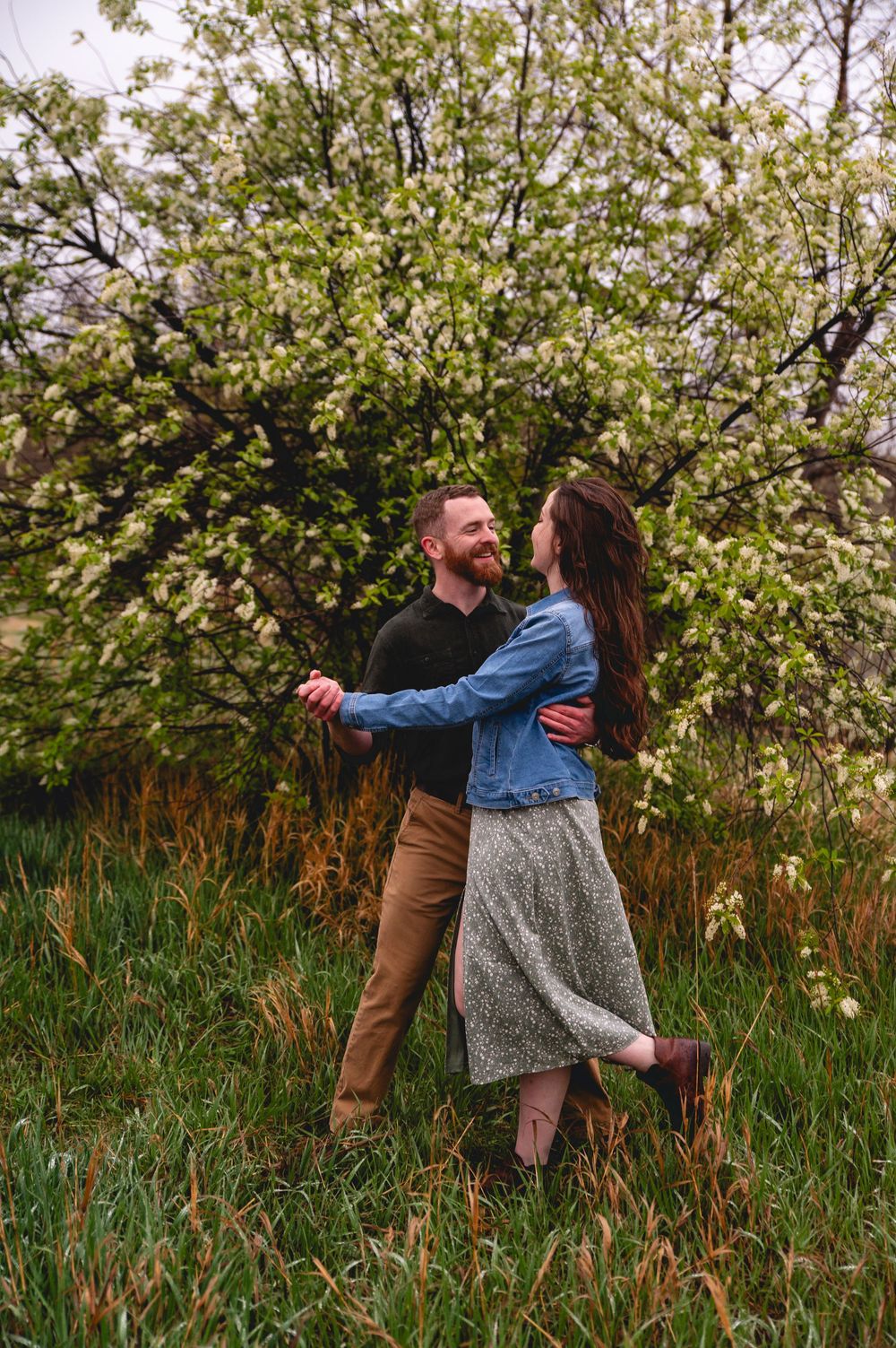 A couple dances together in a blooming spring garden with white flowering trees and tall grass.