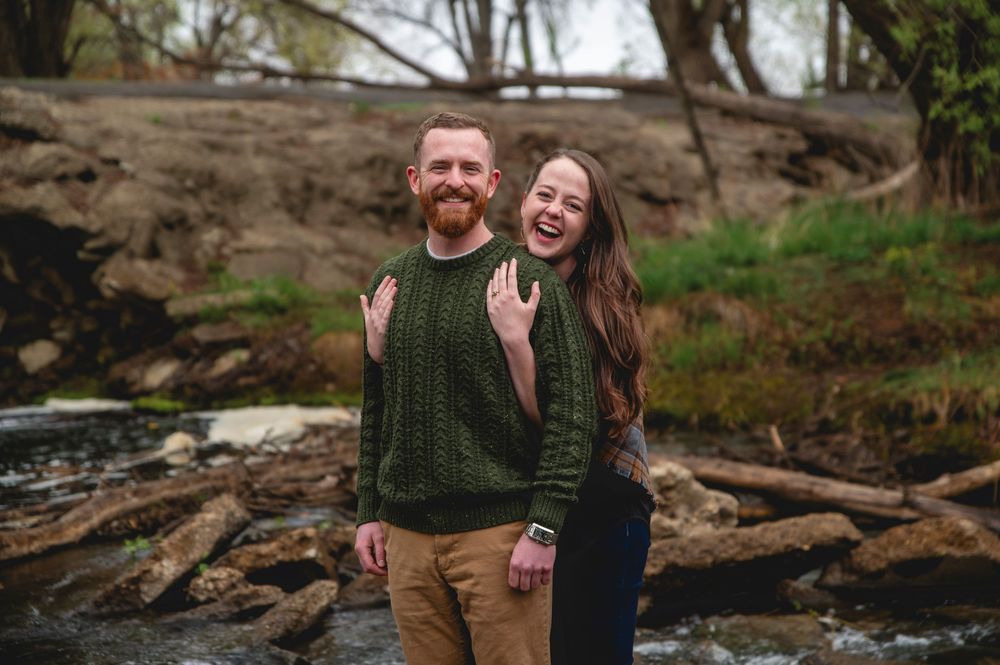 A couple in matching dark green sweaters laughs together while standing by a rocky stream on an autumn day.