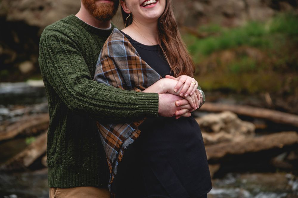 Couple embraces by a stream wearing cozy autumn sweaters and plaid in an outdoor natural setting.