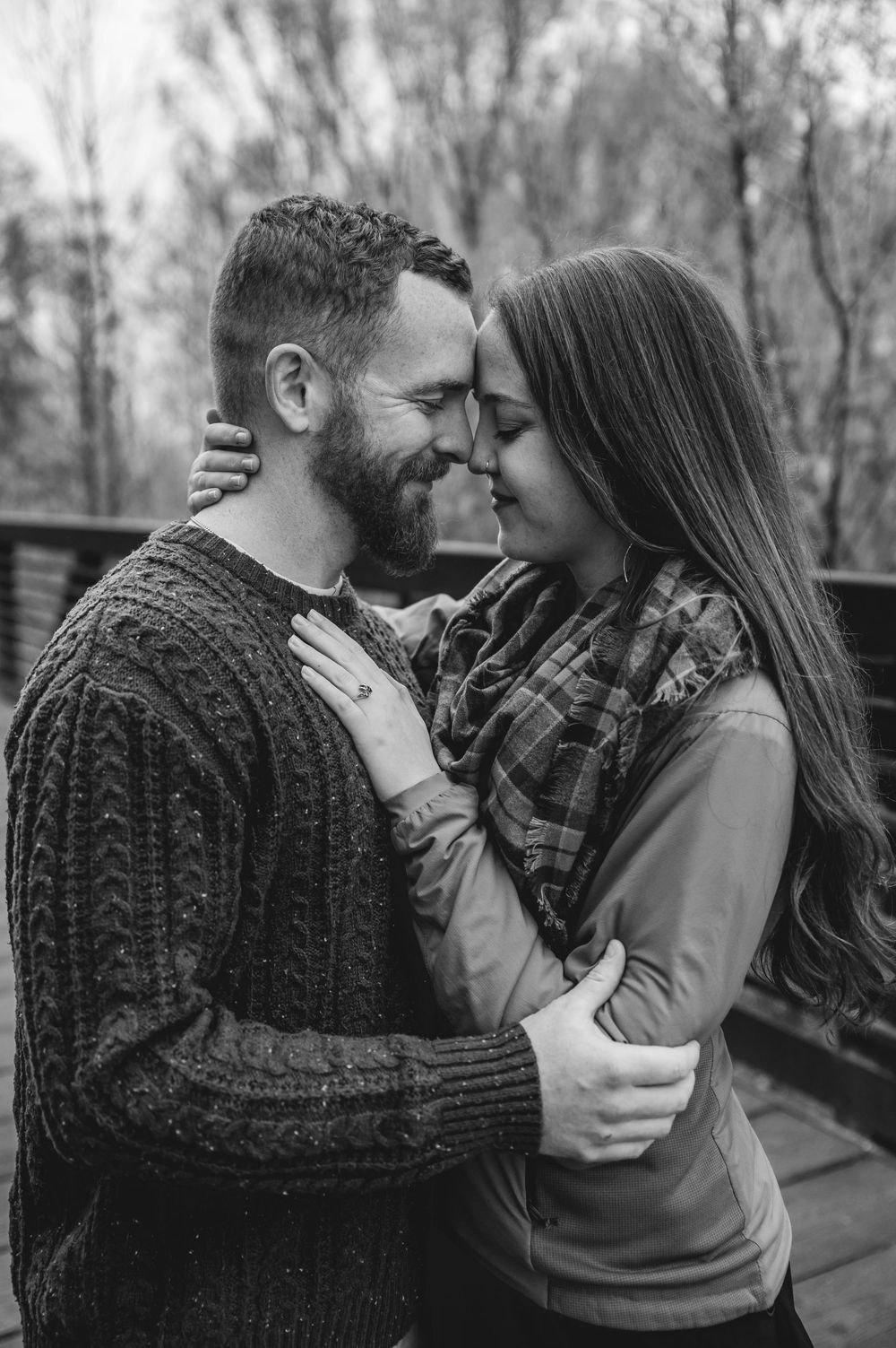A romantic black and white portrait of a couple embracing outdoors, with the backdrop of trees in soft focus.
