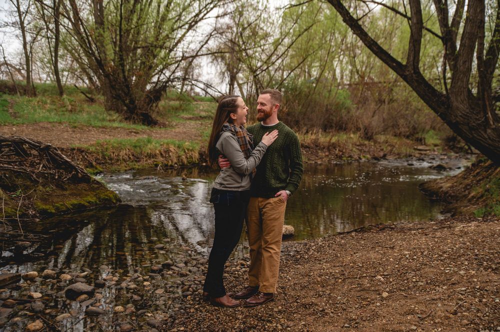A couple embraces by a peaceful stream on an autumn day with willow trees in the background.
