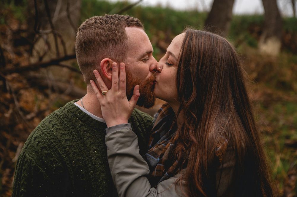 A couple shares a romantic kiss while standing in a wooded area during autumn.