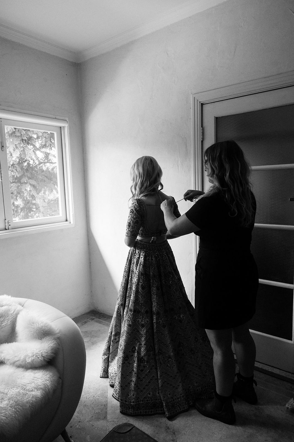 Black and white photo shows two silhouettes in a room, one wearing a long dress while being helped with garment adjustments.