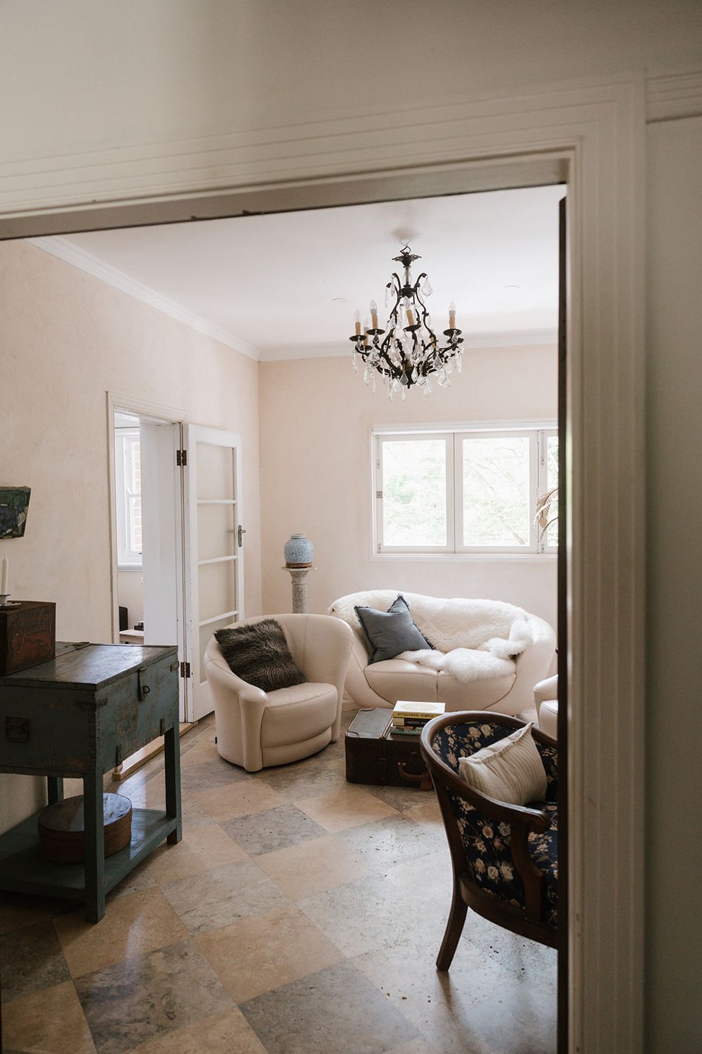 An elegant living room with a cream sofa, black accent furniture, chandelier and stone flooring viewed through a doorway.