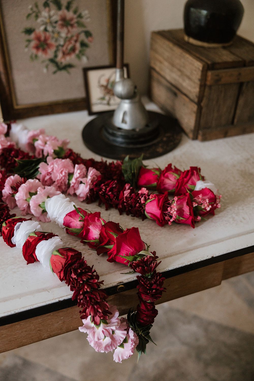 A garland of pink and red dried flowers drapes over a table with vintage decor including wooden crates and floral artwork.
