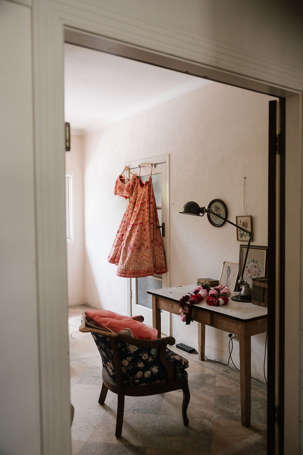 A vintage coral-colored dress hangs on a mannequin beside an antique chair and side table with framed photos.