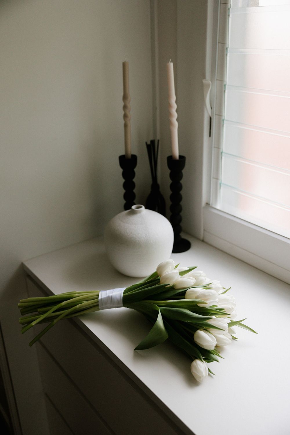 White tulips tied with ribbon rest on windowsill beside black candlesticks and white ceramic vase in moody lighting.