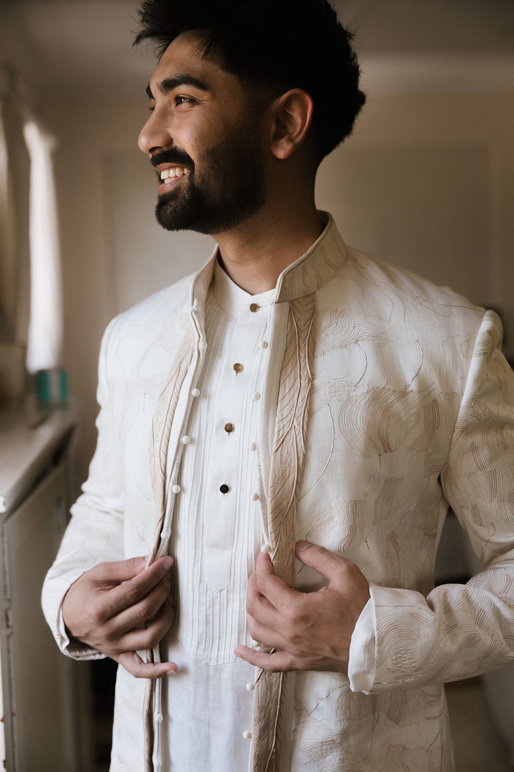 Smiling person wearing traditional white embroidered Indian sherwani suit with button details stands in elegant pose.