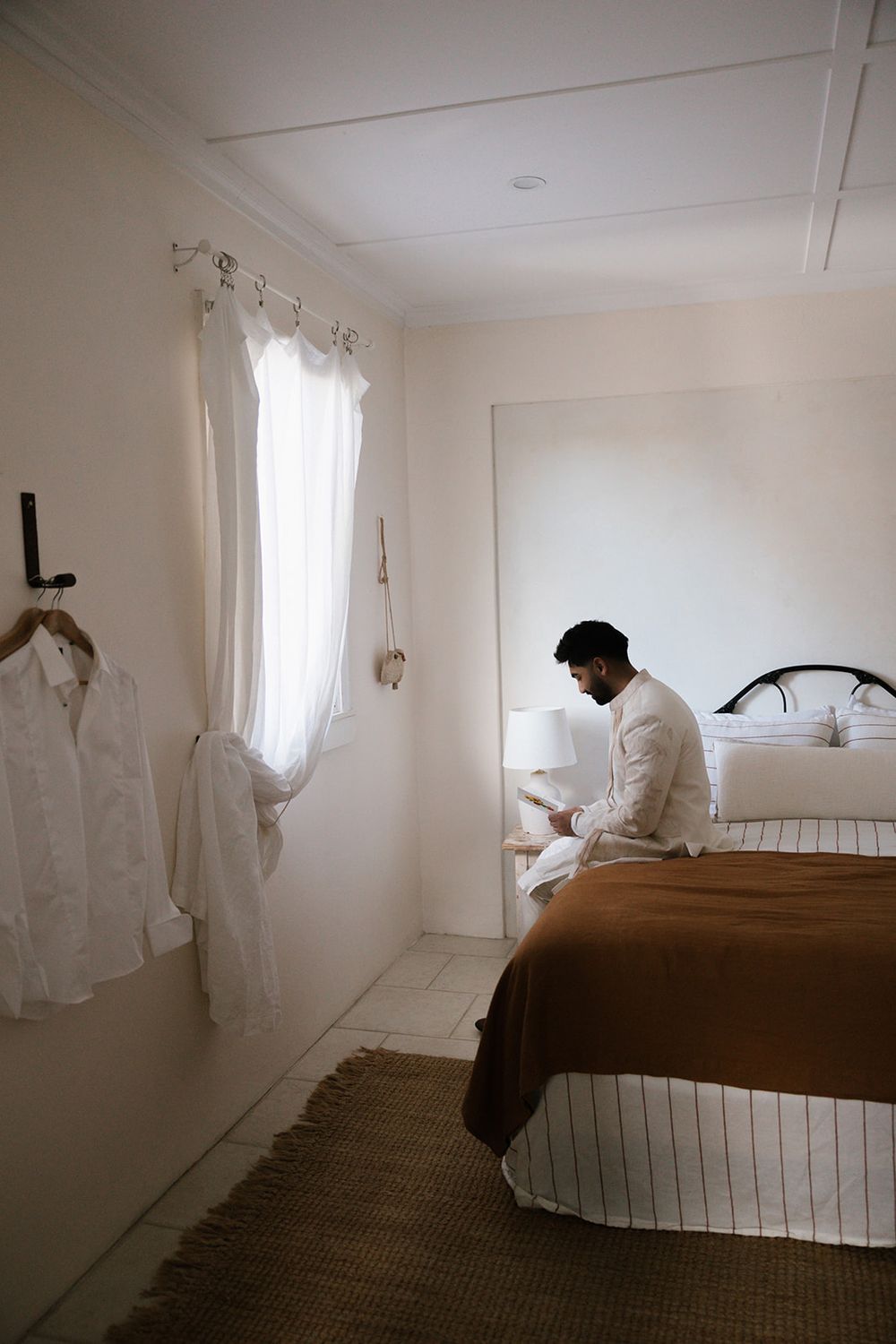 A person in white clothing sits on the edge of a bed in a serene, light-filled bedroom.