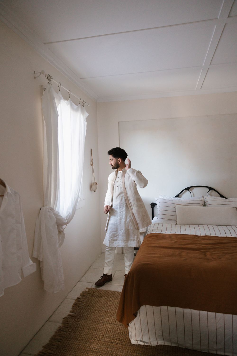 Person in traditional white attire stands in a minimal bedroom with jute rug and brown bedding.