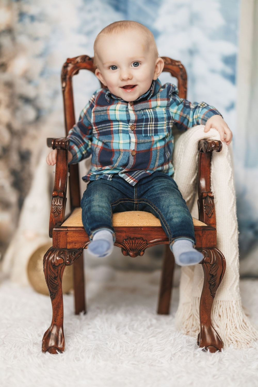 A small child sits in an ornate antique wooden chair with claw feet, wearing a plaid shirt and jeans on a light background.