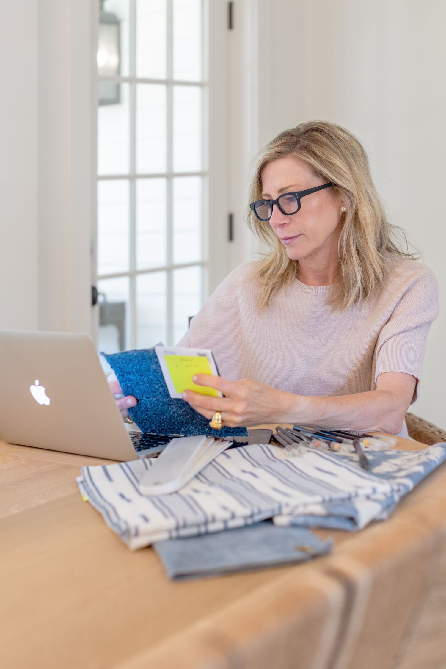 Woman wearing glasses works at a laptop while reviewing notes, seated in a bright room with French doors in the background.