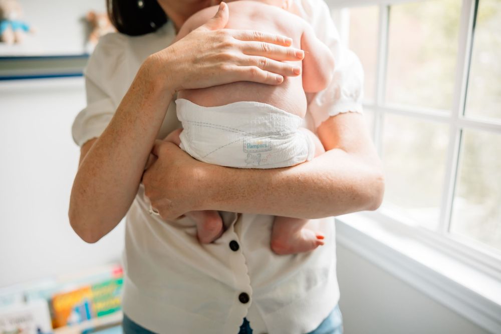 A close-up view of someone in a white shirt holding a sleeping baby near a sunlit window.