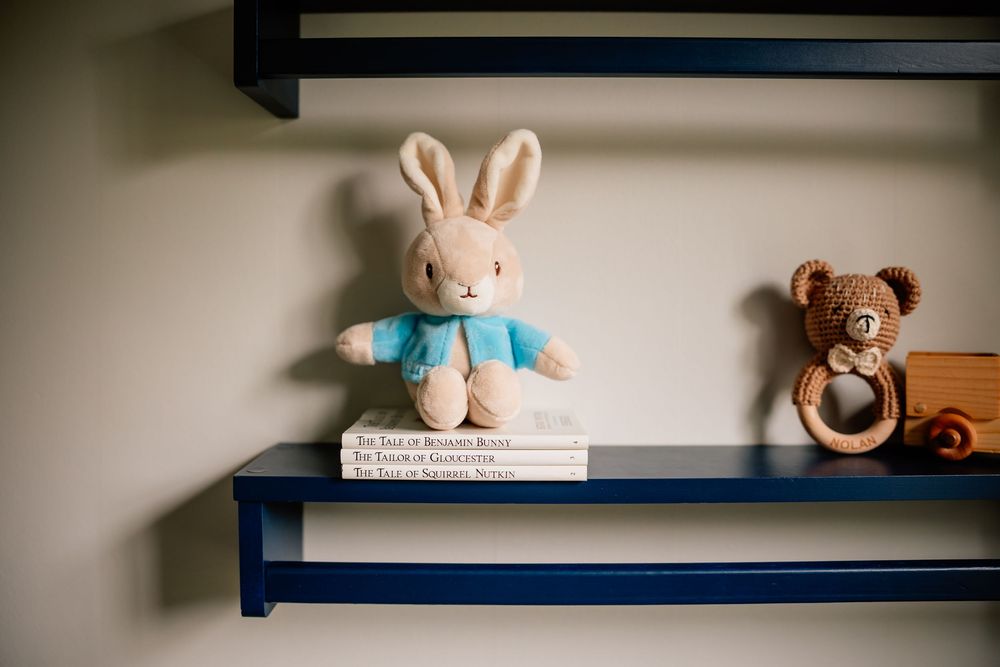 A plush bunny in a blue shirt and brown teddy bear sit on navy blue floating shelves in a nursery.