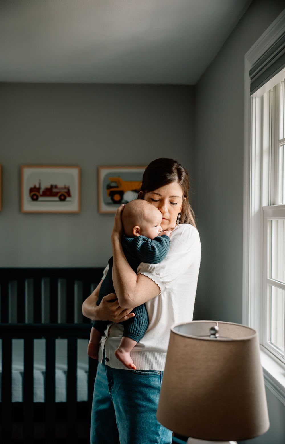 A tender moment by a nursery window showing a parent embracing a baby in a dimly lit room with framed artwork.