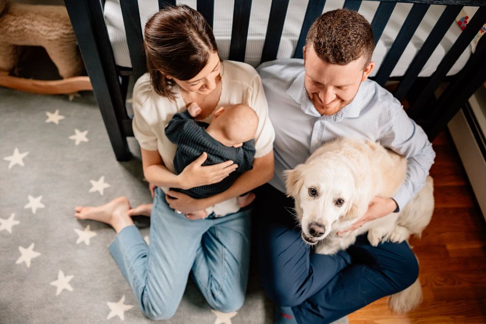 A couple sits on a star-patterned rug with their white dog and newborn baby at home.