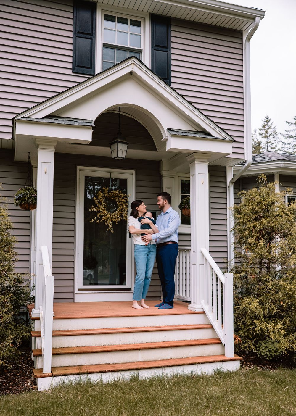 A family stands together on the front porch steps of their tan colonial style home with black shutters.