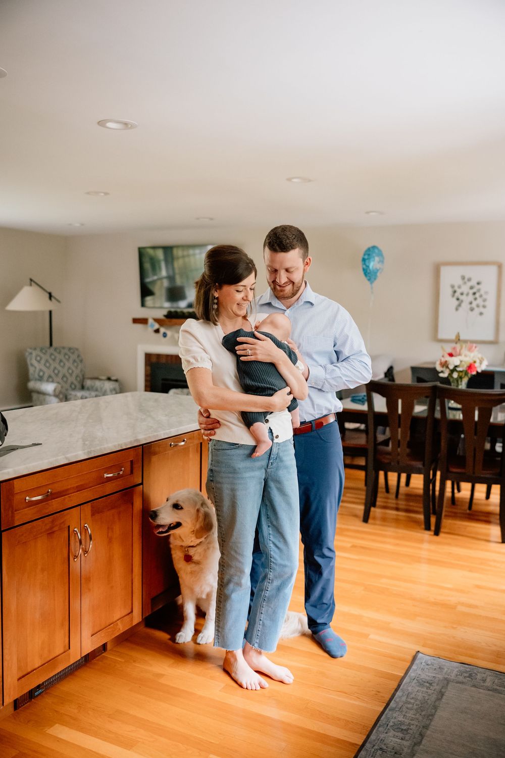 Parents standing in kitchen with infant while golden retriever dog sits beside them on hardwood floor.