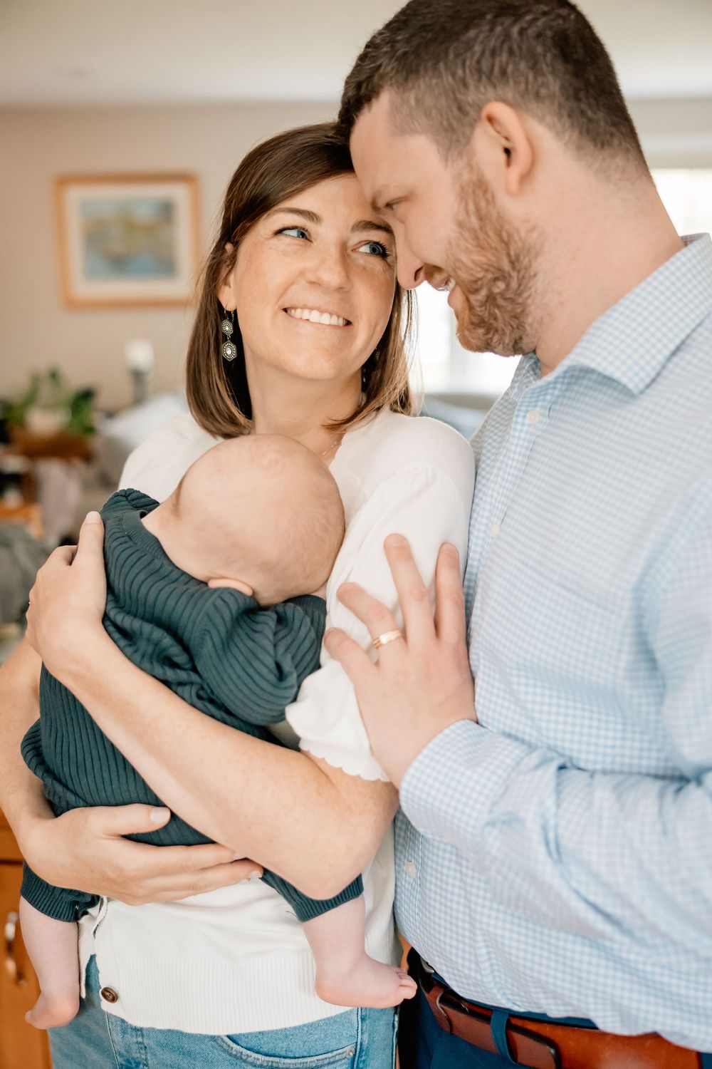 A cozy indoor family moment shows a small group sharing an intimate embrace while holding their baby.