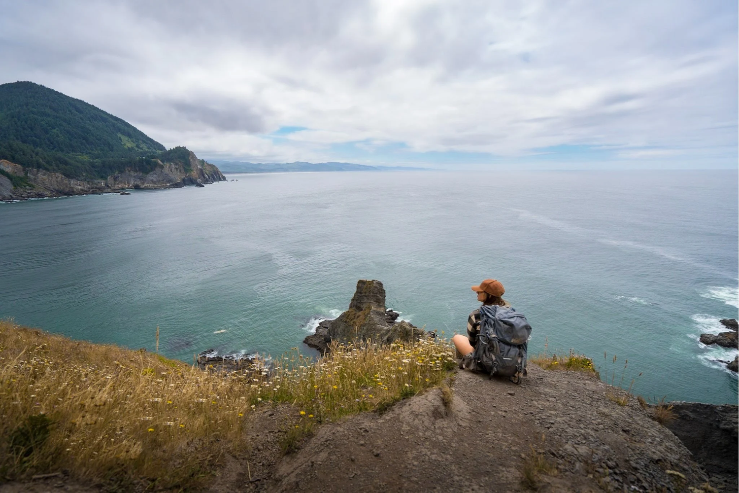 Person sits on cliff edge overlooking dramatic coastal landscape with rugged sea stacks and misty ocean views.