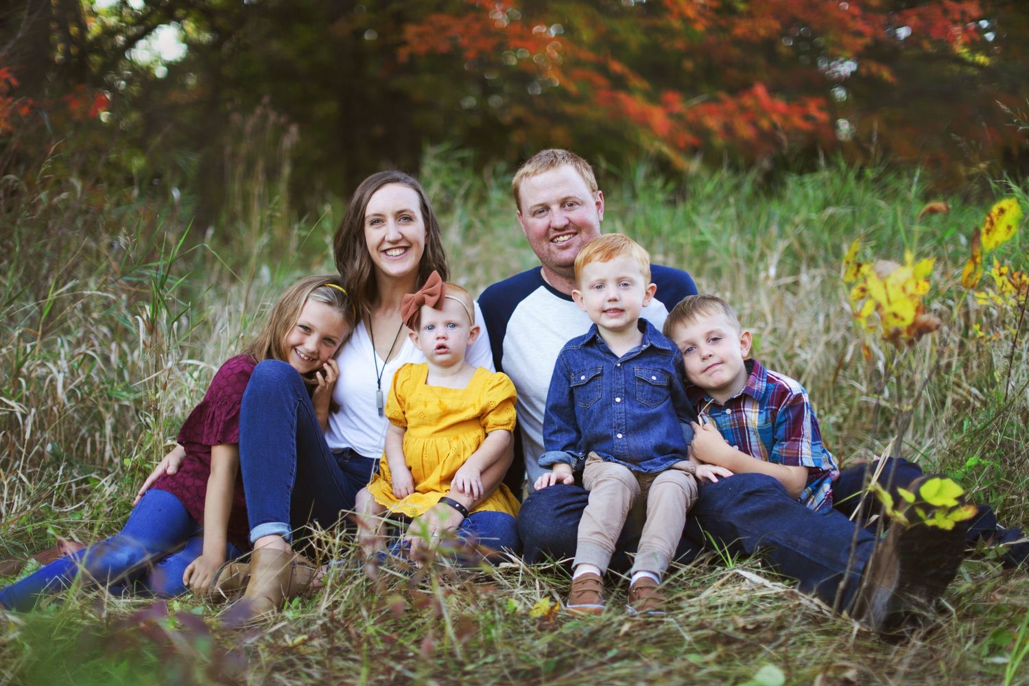 A family group sits together in a fall meadow with red autumn foliage and yellow wildflowers in the background. Duluth Family photographer