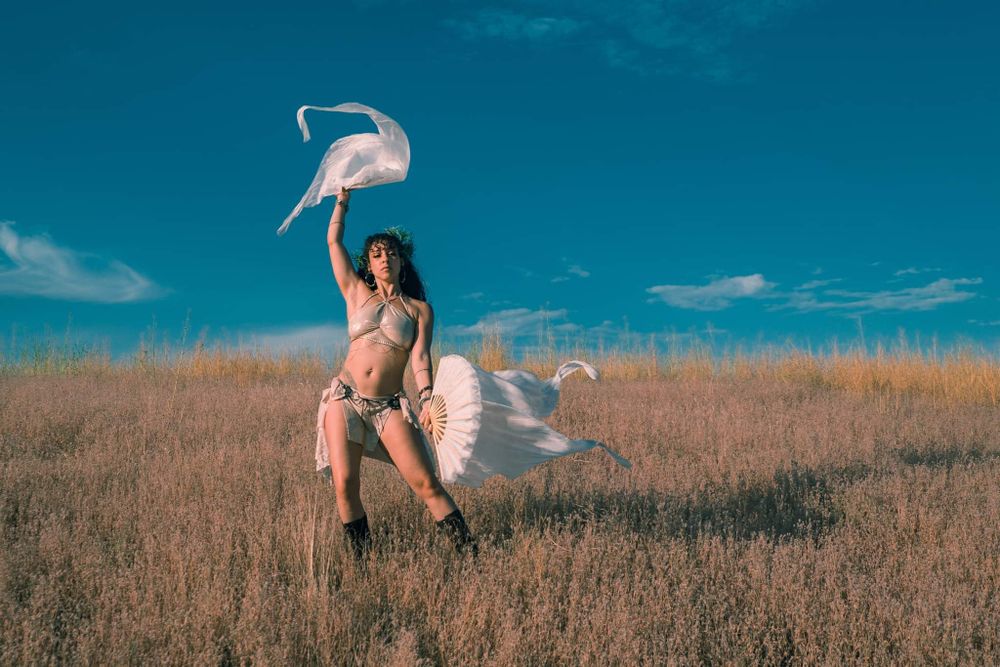Dancing figure in field with white flowing fabric against vibrant blue sky.