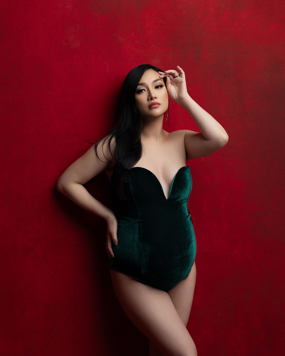 Studio portrait of a woman in an emerald velvet bodysuit posing against a deep red backdrop.