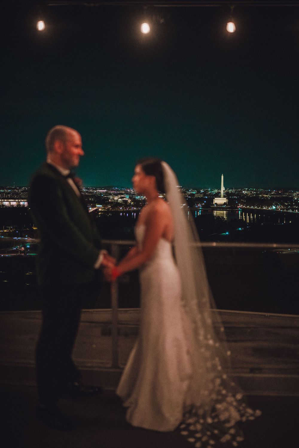A couple in wedding attire stands silhouetted against a the DC skyline at night with string lights overhead.