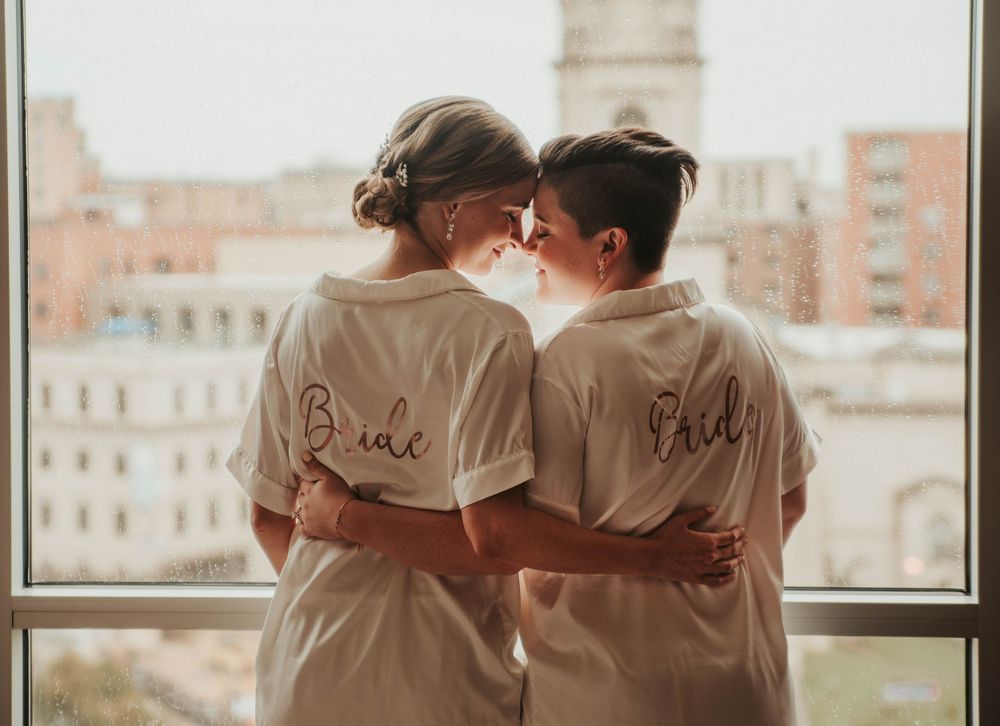 Brides in matching robes labeled 'Bride' embrace while standing near a large window overlooking Washington, DC city buildings.