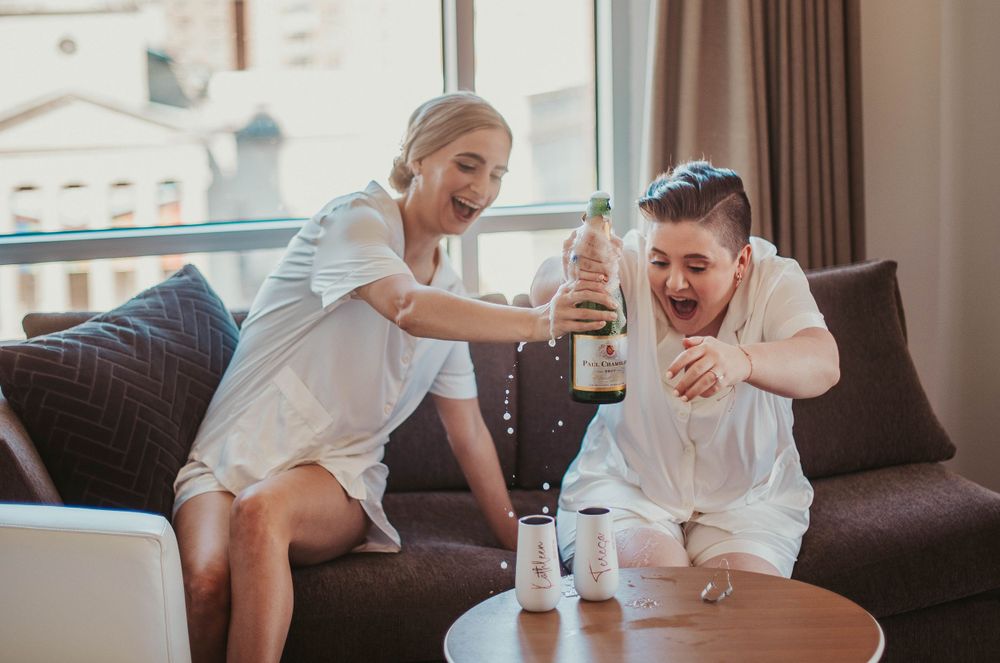 Two brides popping champagne in Washington, DC, liquid dripping down the bottle and surprised expressions on their faces.