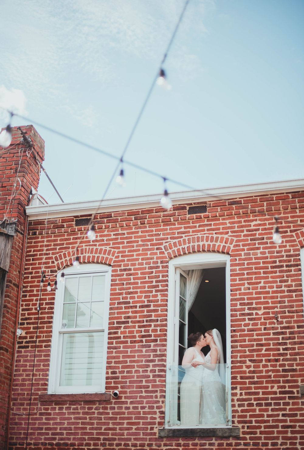 A red brick building exterior with string lights and a window with white shutters and a doorway and two brides kissing in the window in DC.