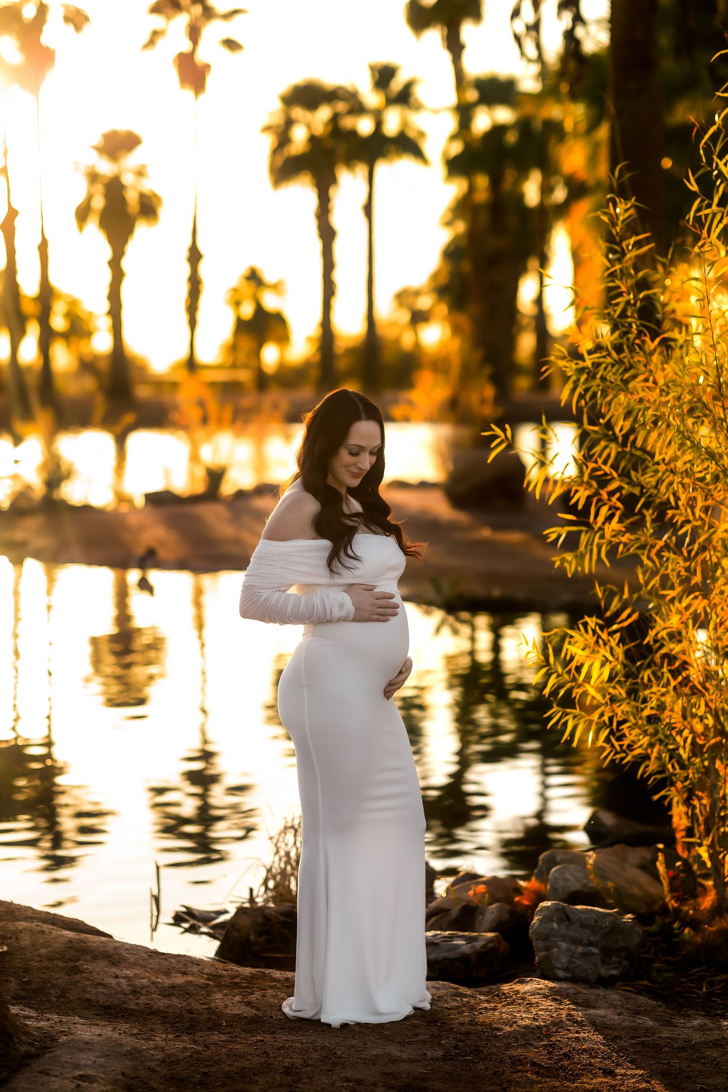 Serene maternity portrait in white dress by reflective water during golden hour at Papago Park in Phoenix.