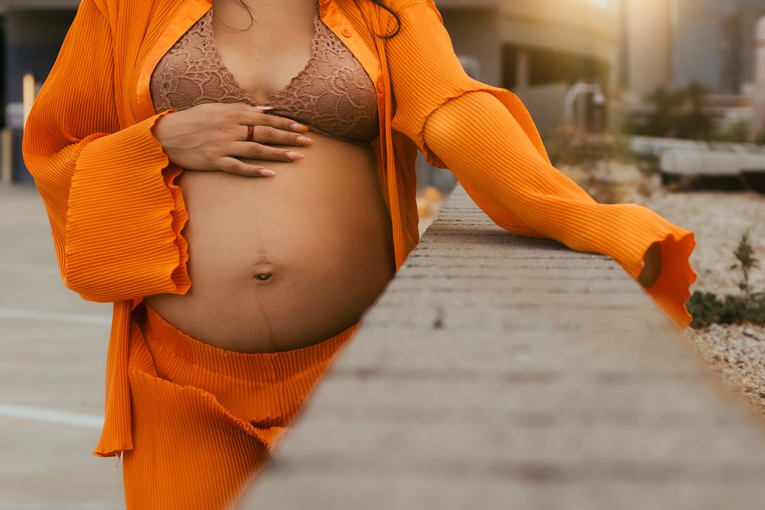 Closeup of pregnant belly with hands cradling bump, wearing orange outfit against city backdrop in Downtown Phoenix.